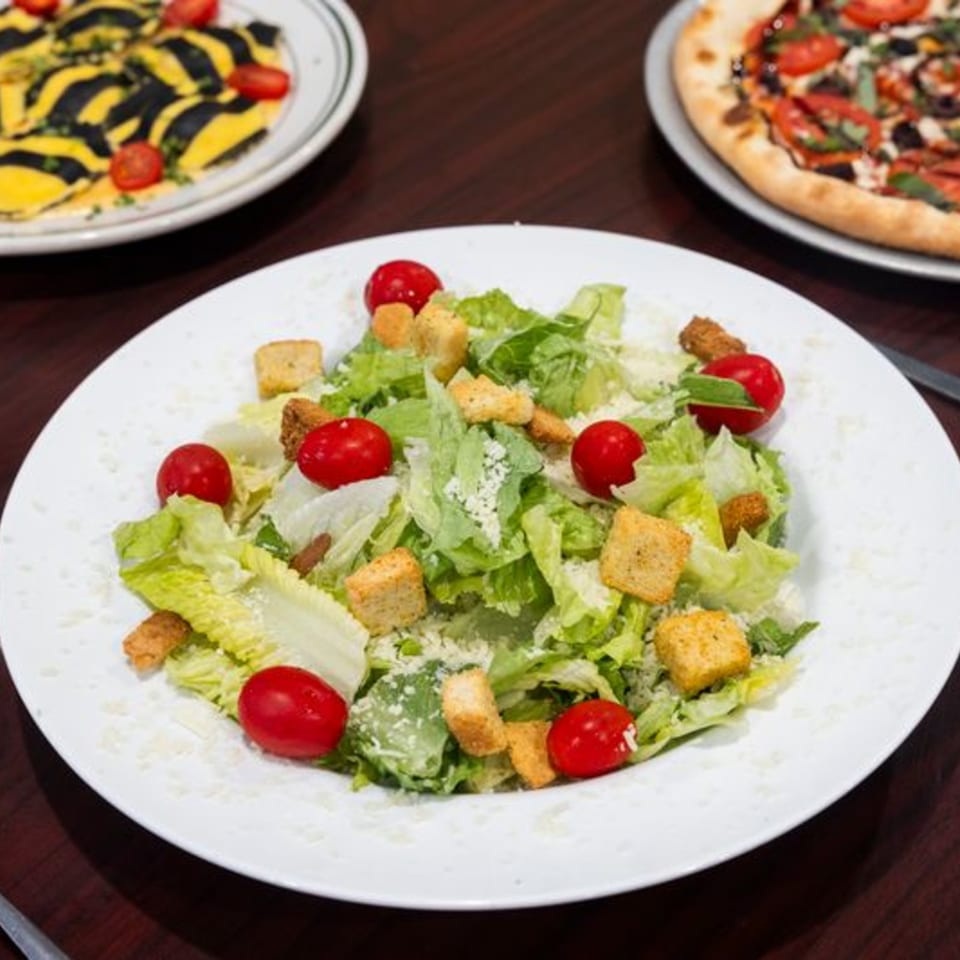 A Caesar salad with cherry tomatoes, croutons, and parmesan cheese, with pasta and pizza in the background.