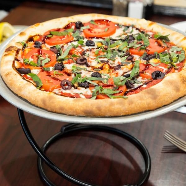 A freshly baked pizza with tomatoes, olives, basil, and balsamic glaze on a metal stand.