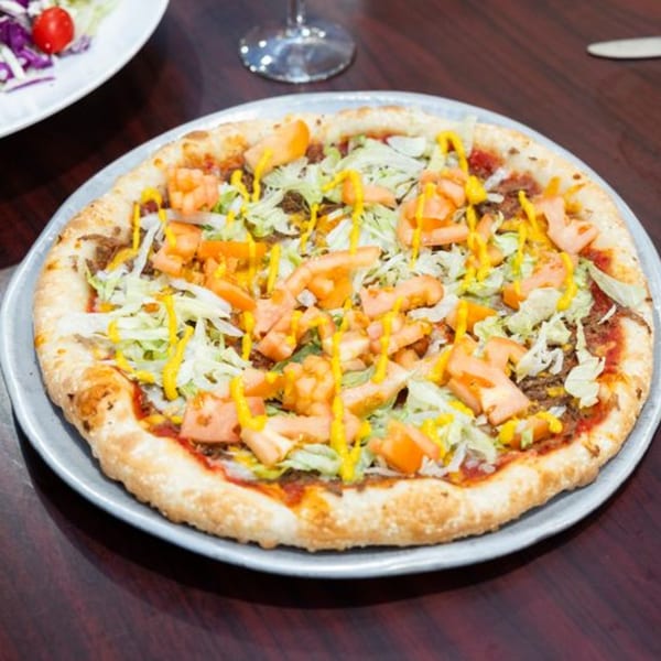 A taco pizza with ground beef, shredded lettuce, diced tomatoes, and yellow mustard drizzle.