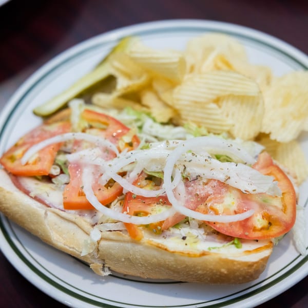 A submarine sandwich with tomatoes, onions, and lettuce, served with potato chips and a pickle.