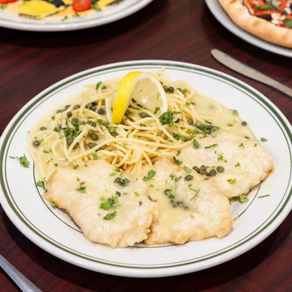 Chicken piccata with spaghetti, capers, parsley, and a lemon wedge on a white plate.